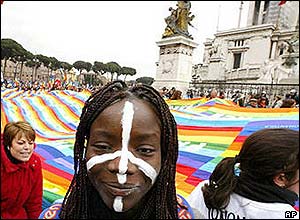 Woman with peace sign painted on her face stands in front of protesters carrying giant peace flag in Rome.